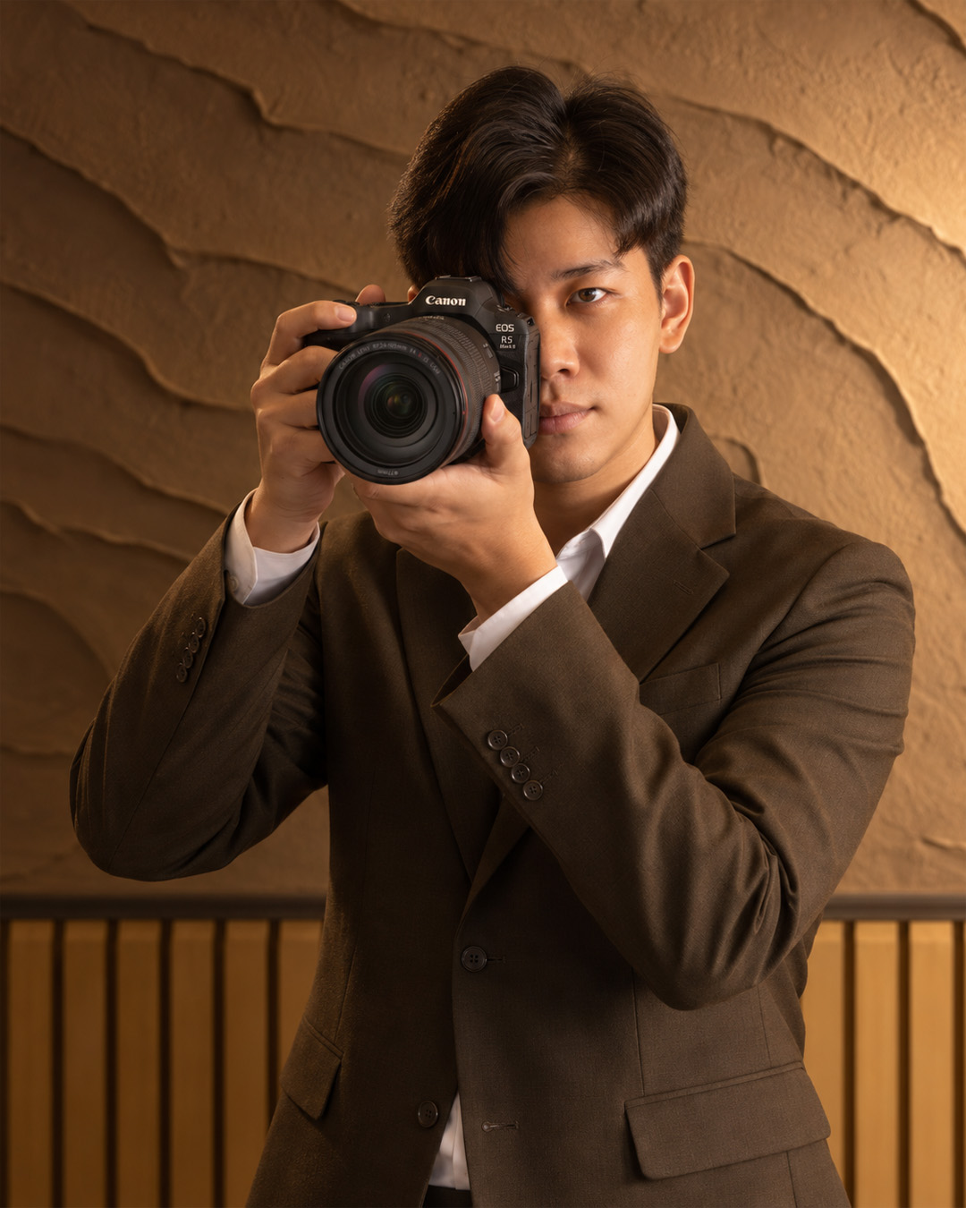 Jatu สวมสูทสีน้ำตาลถือกล้อง Canon ถ่ายภาพ โดยมีผนังโทนอุ่นเป็นฉากหลัง